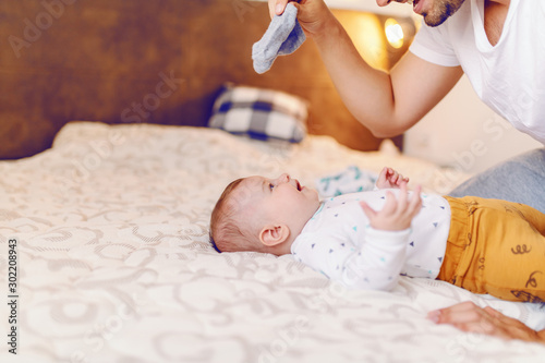 Happy handsome caucasian young dad holding tiny baby socks and preparing to change clothes to his laughing cute 6 months old son. Bedroom interior.
