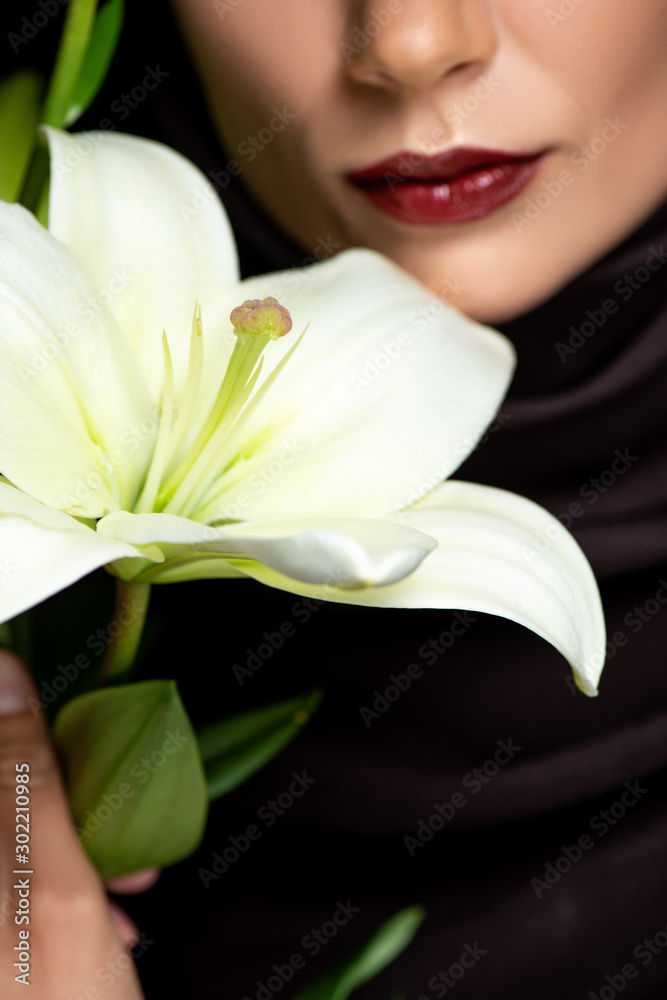 cropped view of beautiful Muslim woman in hijab with red lips holding lily isolated on black cropped view of beautiful Muslim woman in hijab with red lips holding lily isolated on black