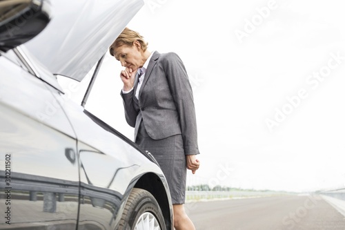 Worried businesswoman looking at breakdown car on road against sky © moodboard