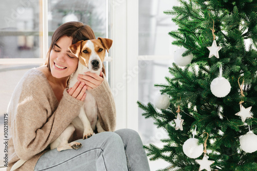 Horizontal shot of caring smiling woman hugs puppy with love, enjoys cozy atmopshere and lovely moment, smiles pleasantly, decorate New Year tree together, celebrate favourite holiday at home photo