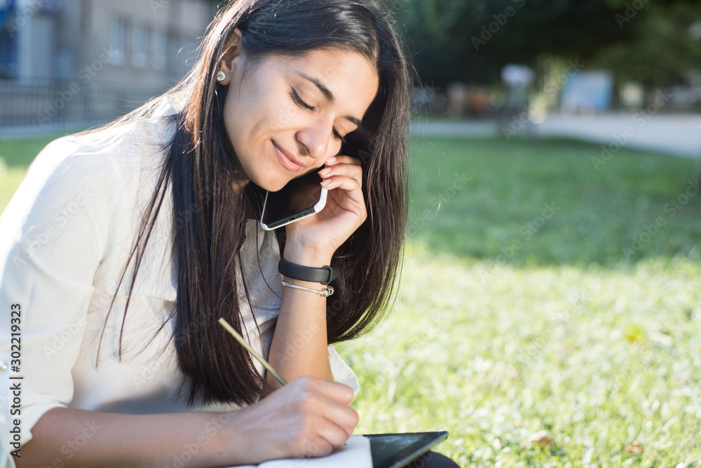 young indian girl Portrait of a beautiful young indian girl. Student, business woman, sitting  on the green grass, talking on the phone and taking notes in a notebook  Stock 写真 | Adobe Stock