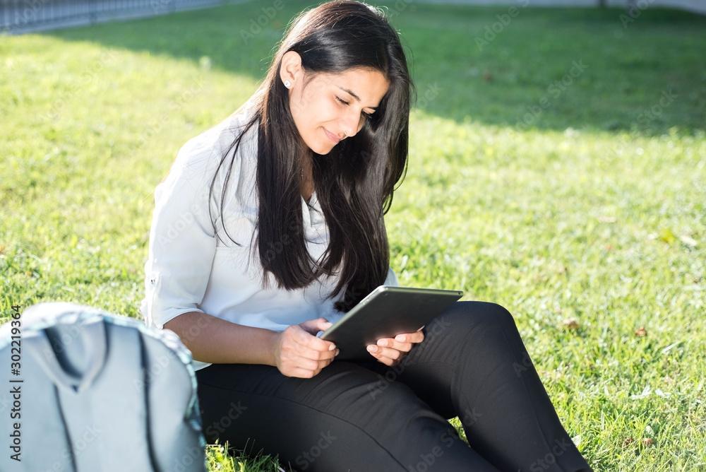 portrait of a beautiful indian girl. Student sitting on green grass, with backpack, smiling, holding tablet in hands portrait of a beautiful indian girl. Student sitting on green grass, with backpack, smiling, holding tablet in hands