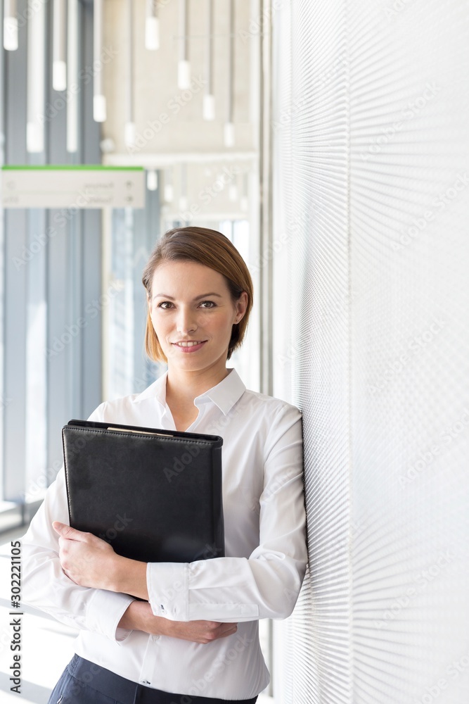 Businesswoman leaning on wall while holding black leather document in office Businesswoman leaning on wall while holding black leather document in office