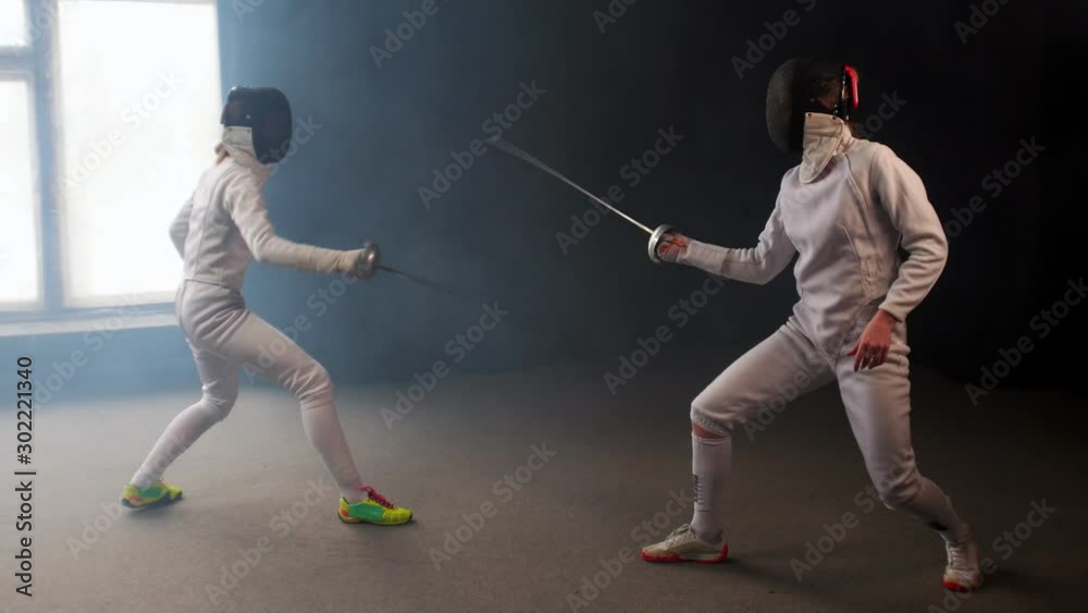 Two young women having an intense training in a fencing duel in the smoky studio Two young women having an intense training in a fencing duel in the smoky studio