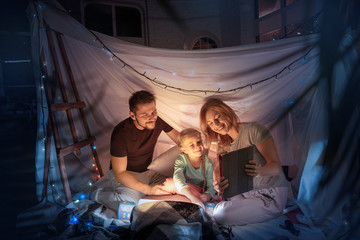 Caucasian family sitting in a teepee, having fun and playing with the flashlight in dark room with toys and pillows. Look happy. Home comfort, family, love, Christmas holidays, storytelling time.