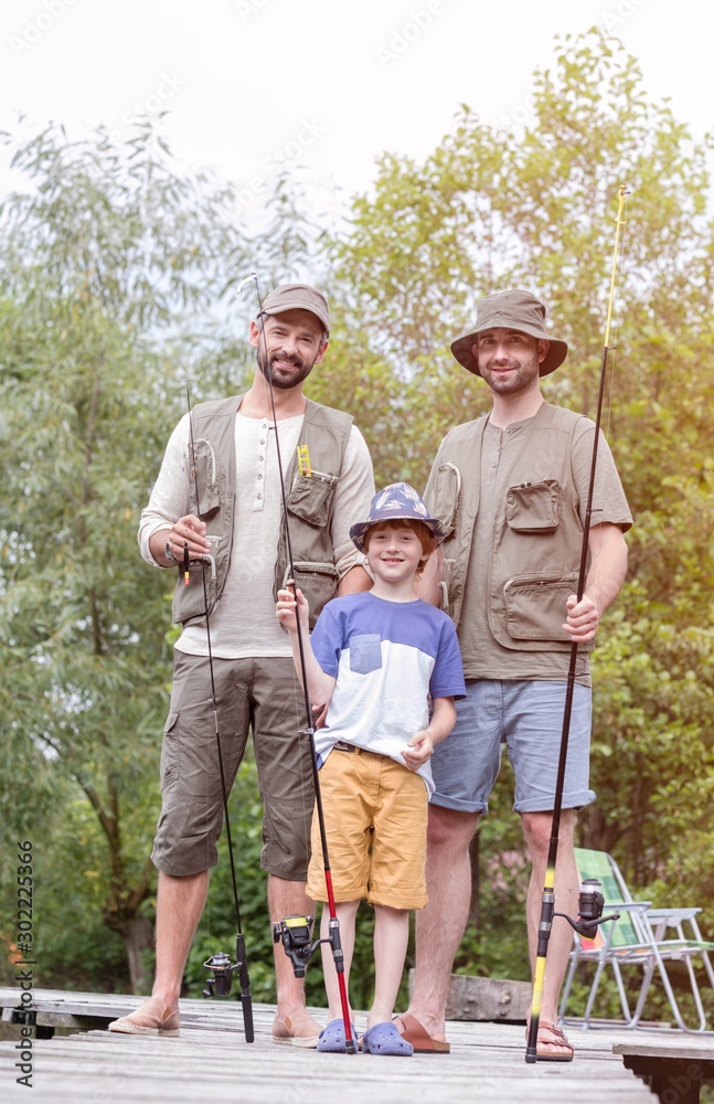 Full length portrait of smiling males standing with fishing rods on pier against trees Full length portrait of smiling males standing with fishing rods on pier against trees
