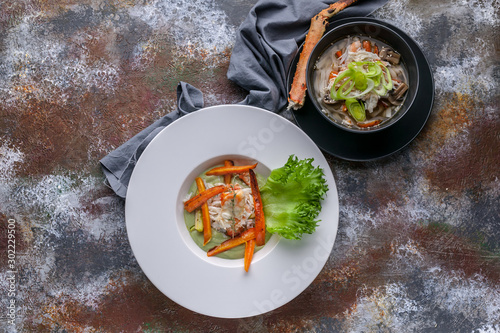 Close-up salad with snow crab meat, soup with snow crab meat, udon noodles and vegetables. Serving on a gray napkin and a rusty background. Top view