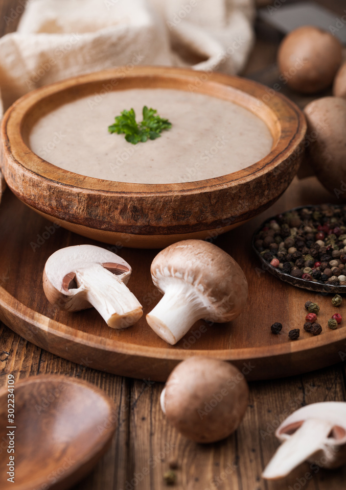Wooden plate of creamy chestnut champignon mushroom soup with wooden spoon, pepper and kitchen cloth on wooden background.
