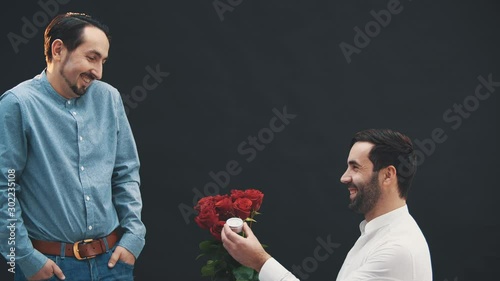 Man is standing on one knee with ring and roses in her hands, making proposal to another man. He says yes. They are happy homosexual couple. photo