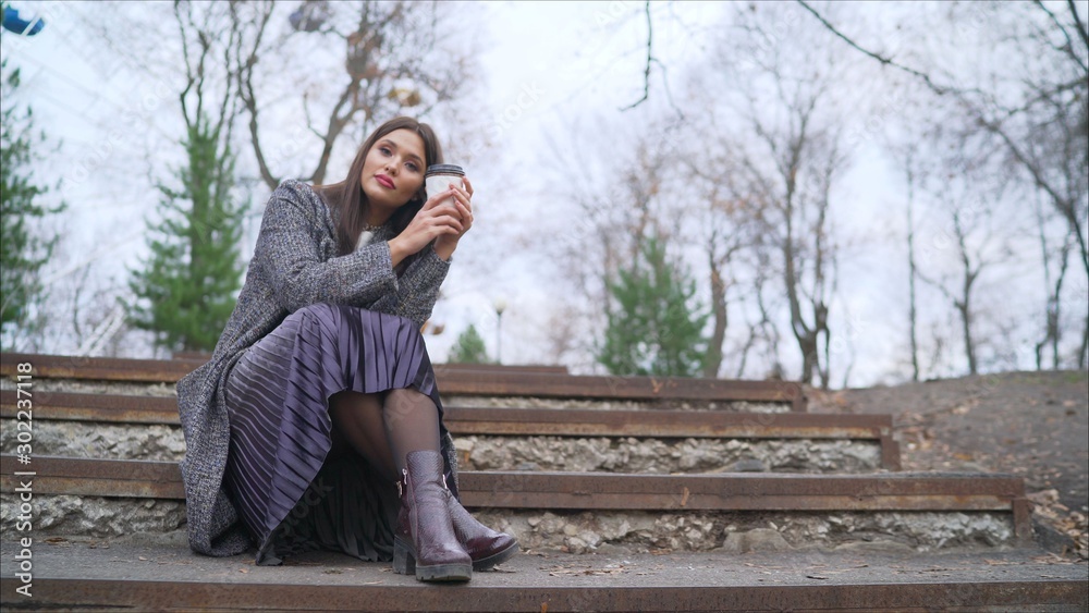 A girl in a coat sits on the stairs and drinks coffee. Portrait of a beautiful girl with long hair in a coat sitting on the stairs. She is looking at coffee.