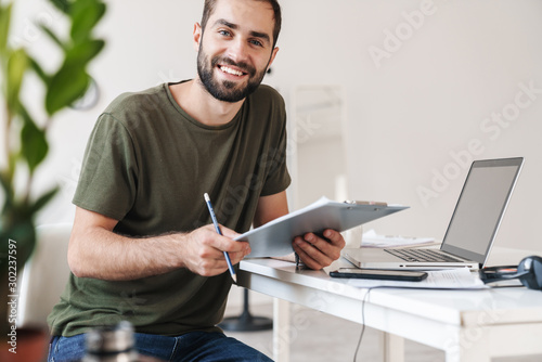 Image of smiling young man using laptop and holding clipboard