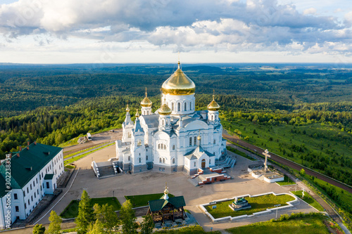 Aerial view of Belogorsky Monastery - russian orthodox church cathedral building with golden domes on hill top among green forest landscape at sunset. Belaya Gora, Perm krai, Russia photo