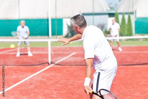 Man swinging racket while playing tennis doubles on red court during summer weekend © moodboard