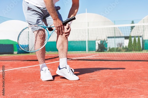 Low section of mature man holding tennis racket while suffering from knee pain on red tennis court during summer © moodboard