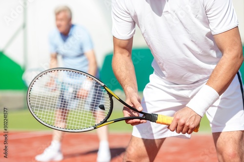Midsection of man standing with tennis racket against friend playing doubles match on court © moodboard