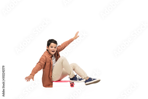 cheerful boy sitting on penny board and balancing with outstretched hands on white background photo