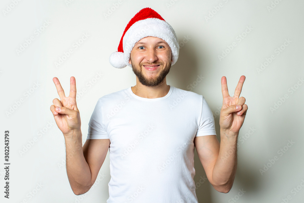 A young man in a Santa Claus hat makes a gesture with his hands on a white background. Christmas concept. Gesture of victory, super A young man in a Santa Claus hat makes a gesture with his hands on a white background. Christmas concept. Gesture of victory, super