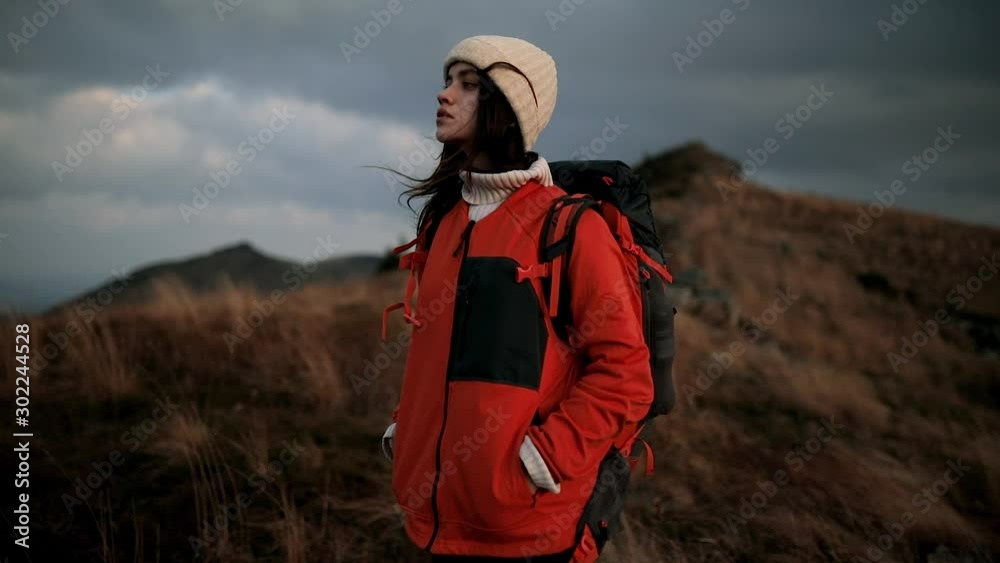 Beautiful dark-haired hiker girl with a backpack stands on top of a mountain and looks at the scenery around. Young woman feels harmony and unity with nature Beautiful dark-haired hiker girl with a backpack stands on top of a mountain and looks at the scenery around. Young woman feels harmony and unity with nature