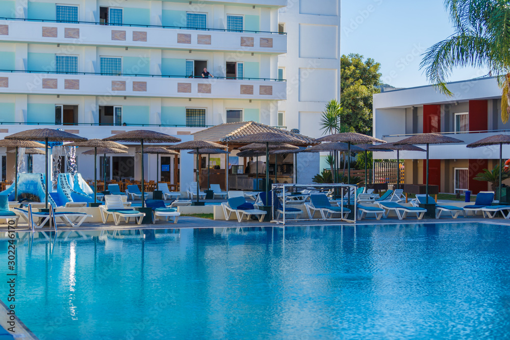 Fototapeta premium Beautiful tropical beach front hotel resort with swimming pool, sun-loungers and palm trees during warm sunny day, paradise destination for vacations