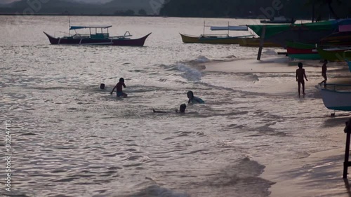 Silhouettes of happy Filipino children playing on the beach at sunset. They jump into the water with a run, swim, and swim amid the swaying boat, it's getting late