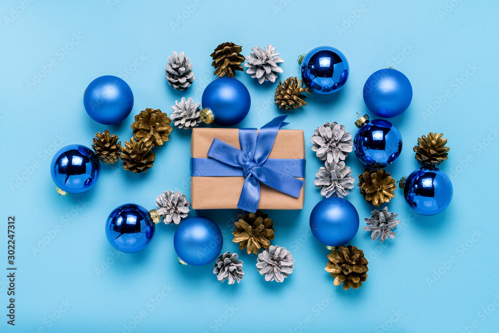Gift box with blue ribbon, Christmas-tree decorations, balls and white and gold pine cones on a blue background. Concept of Merry Christmas and Happy New Year. Minimalism. Flat lay, top view Gift box with blue ribbon, Christmas-tree decorations, balls and white and gold pine cones on a blue background. Concept of Merry Christmas and Happy New Year. Minimalism. Flat lay, top view