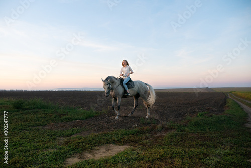 Young blonde girl riding on a horse on the field during sunset