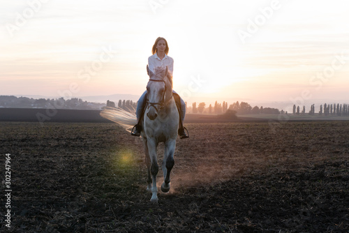 Young blonde girl riding on a horse on the field during sunset © alex_marina