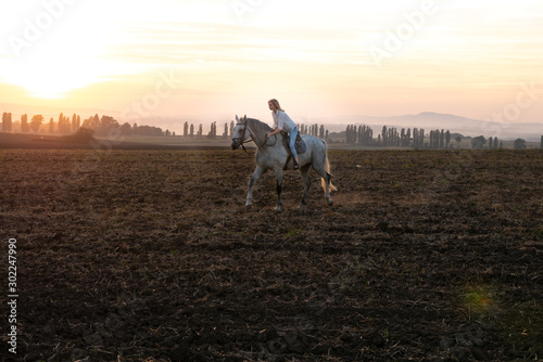 Young blonde girl riding on a horse on the field during sunset