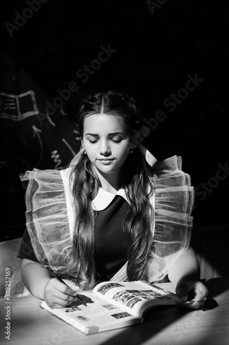 Black and white photo. A schoolgirl in uniform sits at her desk and reads a book. Education, high school, college. Beauty, fashion. A young and beautiful schoolgirl is wearing a traditional uniform
