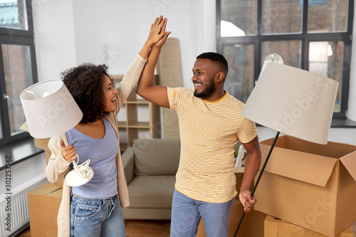 moving, people, repair and real estate concept - happy african american couple with lamps packing stuff into cardboard boxes at home and making high five