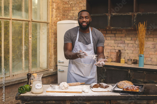 Baking process. Cheerful dark skinned African American male wears apron, kneads dough with great enthusiasm, being content to recieve praise from chef for endeavour and hard work in restaurant.