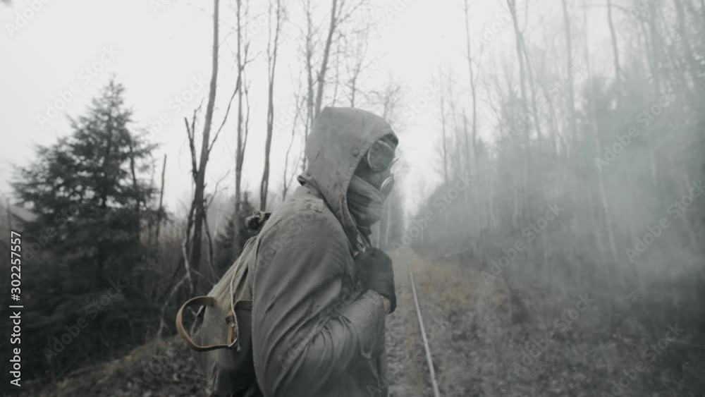 Human in gas mask and military clothes walking on old railway in clouds of toxic smoke. Lonely male survivor stalker after nuclear or chemical war in empty dead forest. Post apocalyptic world Human in gas mask and military clothes walking on old railway in clouds of toxic smoke. Lonely male survivor stalker after nuclear or chemical war in empty dead forest. Post apocalyptic world