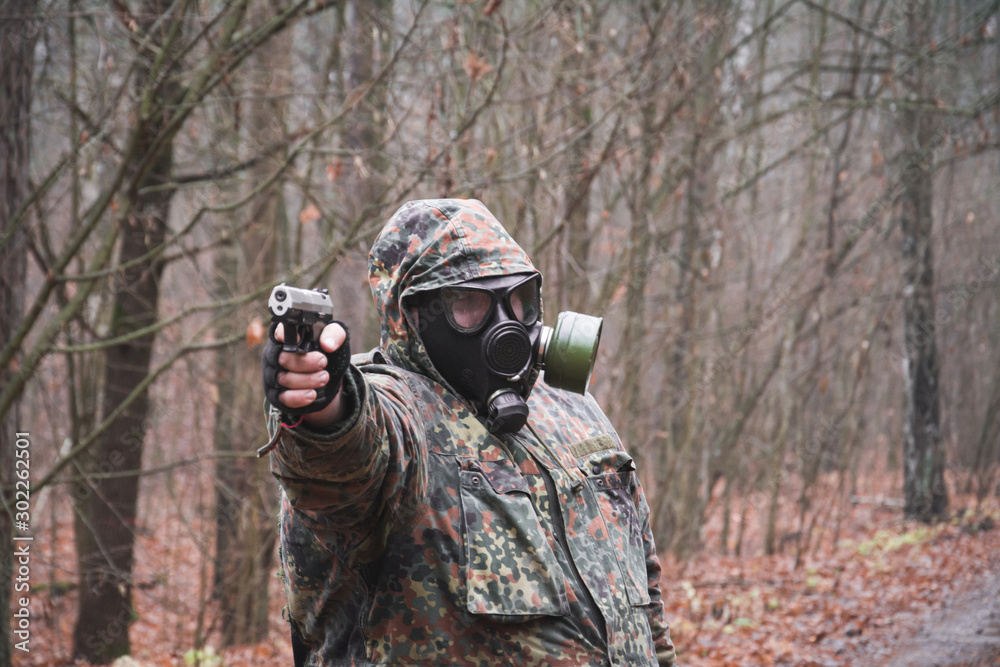 A soldier in camouflage and a gun in his hands in the forest, wearing a gas mask