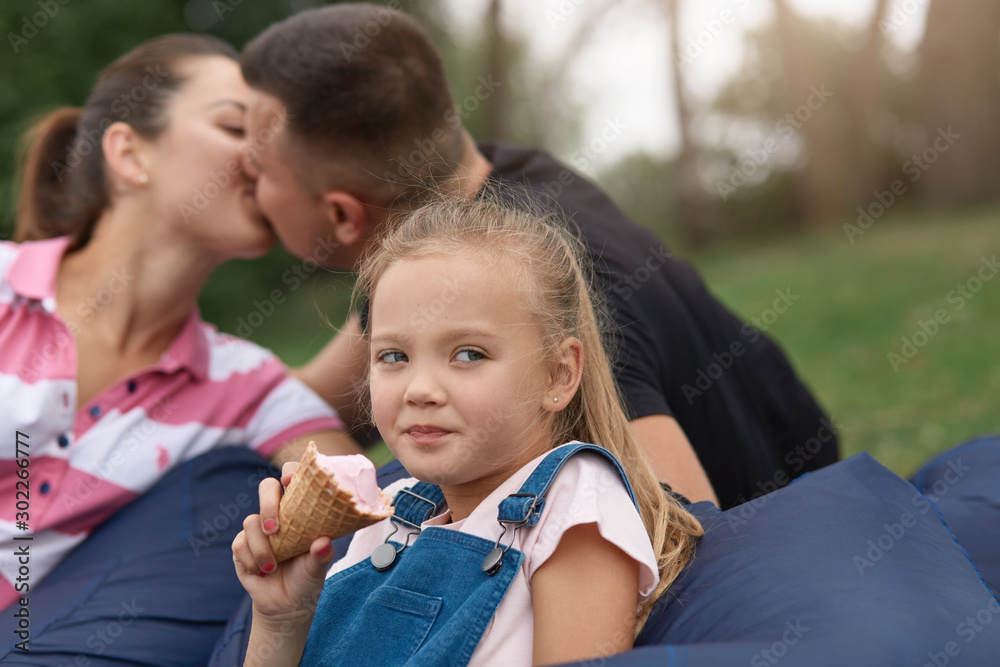 Horizontal shot of happy mother and father kissing while sitting on frameless chair in park with little daughter, cute blonde kid eating ice cream and looks at camera. Happy family have rest in park.