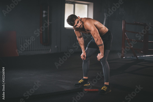 Fototapeta Naklejka Na Ścianę i Meble -  Charismatic guy in sportswear bending with her hands on knees at the gym. Fitness man looking tired after intense crossfit workout