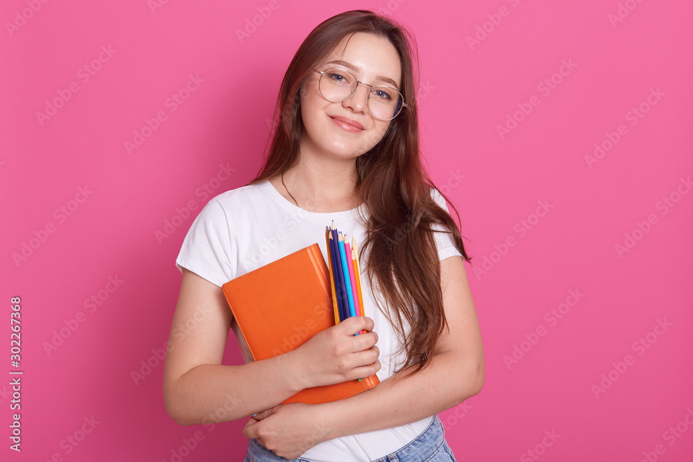 Close up portrait of cute young woman holding textbook and colored pencils,posing in studio isolated over pink background, looking smiling at camera, adorable female wearing white casual t shirt. Close up portrait of cute young woman holding textbook and colored pencils,posing in studio isolated over pink background, looking smiling at camera, adorable female wearing white casual t shirt.