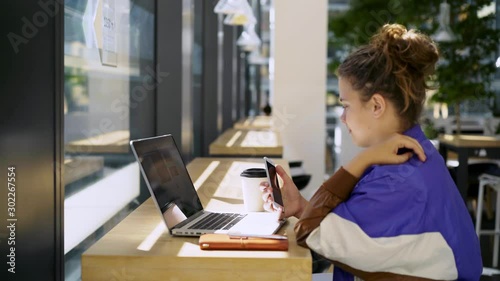 Young woman using laptop at the counter in modern interior against big window. Cropped view female student in trendy wear chatting on smartphone while sitting before notebook connected free wifi