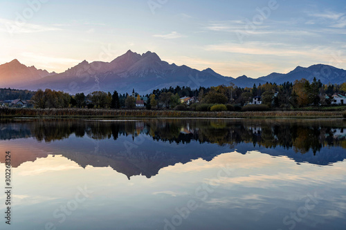 Peaceful scene of beautiful autumn mountain landscape with small settlement, calm lake, colorful trees and high peaks in High Tatras, Slovakia.