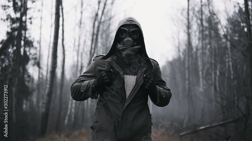 Wandering man in military clothes and gas mask walking on old rail in empty dark forest. Stalker concept, survivor after nuclear or chemical war. Post apocalyptic world. Stedicam following shot