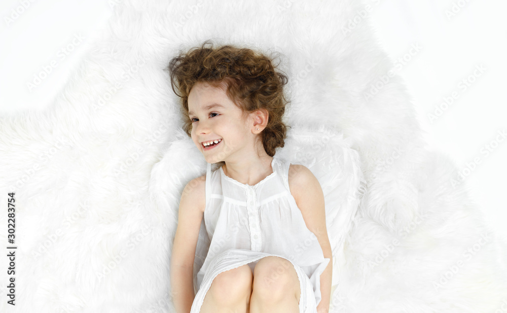 Adorable curly little girl lying down on fur, looking at a side, in white dress and wings, isolated on white background.