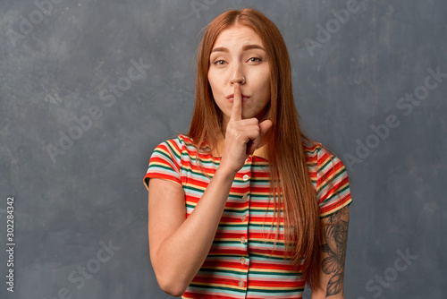 girl holding index finger at lips, asking to keep silence or not tell anyone secret, raising brows. Beautiful young girl with ginger long hair and tattoo on arm, wearing striped top, isolated on wall