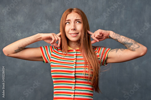 Young handsome girl tries to ignore annoying noise, plugs ears, dressed striped shirt, has displeased expression, isolated on textured background in Studio. refuses to listen something unpleasant.