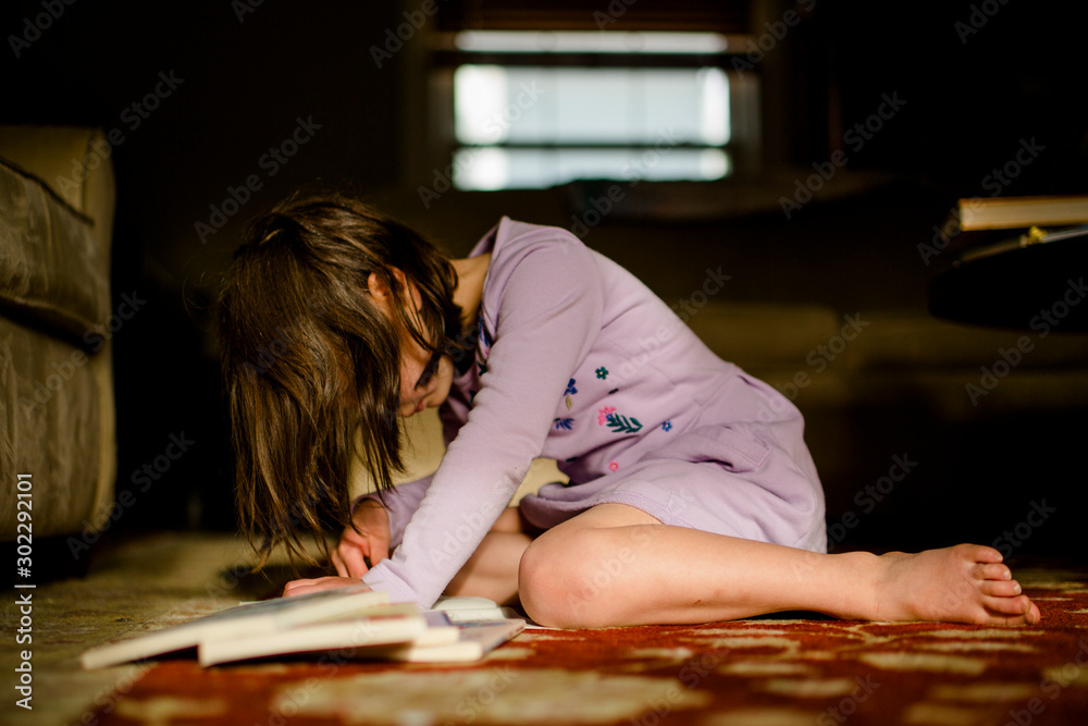 A small child sits on rug in morning light with pile of books reading
