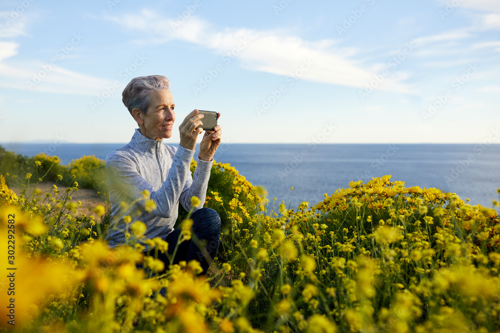 Senior woman photographing through smart phone while crouching by plants by sea against sky Senior woman photographing through smart phone while crouching by plants by sea against sky