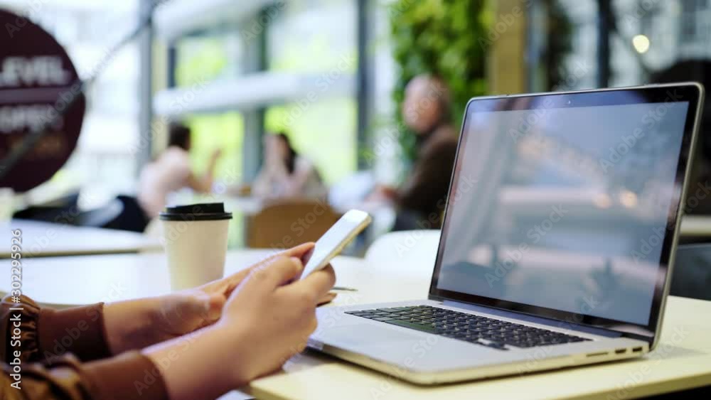 Young woman using laptop at the table in cafeteria against big window. Detail shot female hands messaging on smartphone before notebook. Concept of distant work freelancer in public space