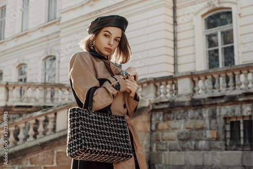 Outdoor autumn fashion portrait of young elegant woman wearing classic beige trench coat, trendy faux leather beret, holding black tweed bag, handbag, posing in street of city. Copy, empty space 