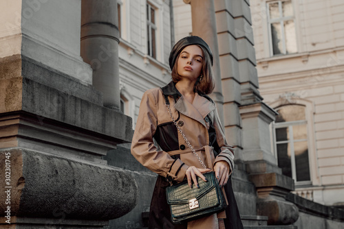 Outdoor autumn fashion portrait of young elegant woman wearing beige black trench coat, beret, with green faux leather textured crocodile shoulder bag, posing in street. Copy, empty space for text