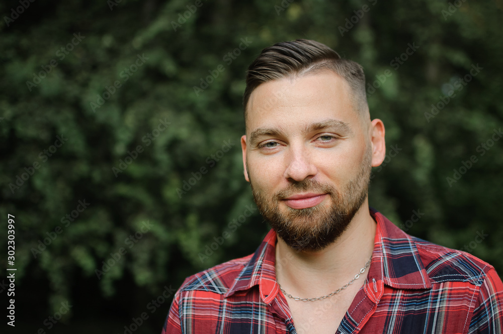 Close up portrait of young bearded man in red shirt looking at the camera with smile in the park on trees background Close up portrait of young bearded man in red shirt looking at the camera with smile in the park on trees background