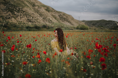 Pensive attractive red haired adult lady in yellow dress with red poppy in hair and red lips looking away while sitting alone in blurred amazing green meadow with red and white flowers against hills under gray cloudy sky