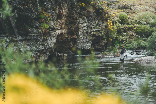 Side view of equipped man harling fish while standing in waders in mountain torrent by cliff and forest photo